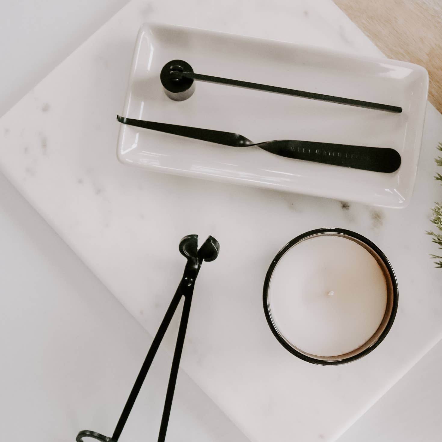 A styled flat-lay of a black metal jewelry and makeup tool set on a white marble tray, alongside a candle and green foliage on a light wood surface.