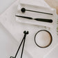 A styled flat-lay of a black metal jewelry and makeup tool set on a white marble tray, alongside a candle and green foliage on a light wood surface.