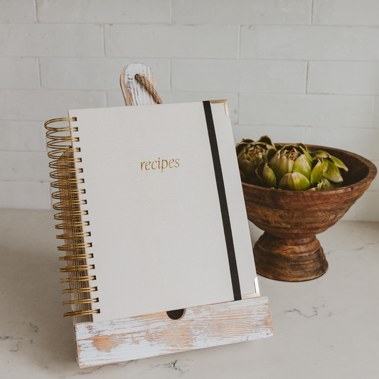 Neutral hardcover heirloom recipe book with gold spiral binding and tabbed recipe sections displayed in a modern kitchen.