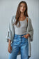 Woman wearing a gray cardigan, white tank top, and blue jeans against a plain background