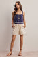 Woman wearing a blue embroidered top and beige shorts on a plain background