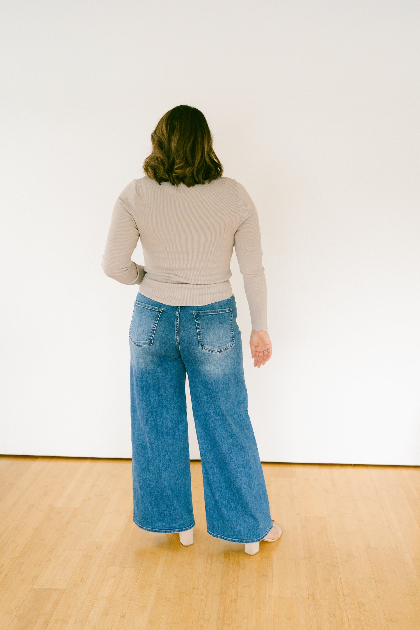 A woman viewed from behind wearing a beige long-sleeve top and wide-leg blue denim jeans, standing on a light wood floor against a plain white wall.