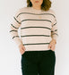 A woman stands in a minimalist room with a white wall and light wood floor, wearing a cream knit sweater with thin black horizontal stripes and dark wide-leg pants.